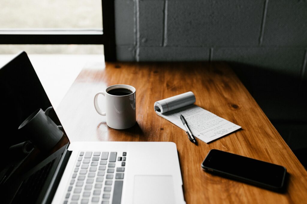 A simple desk setup with a laptop, coffee, notebook, and pen, reflecting a focused work environment for freelancers aiming to avoid scams while working online.